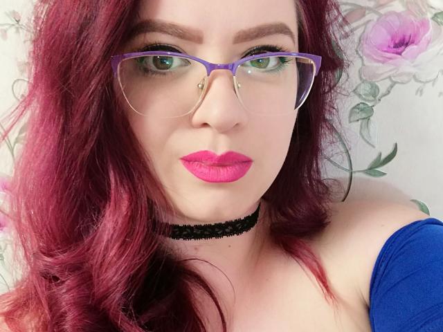 AnaisGrosSeinss, com cabelo ruivo, óculos roxos, batom rosa, colar preto e blusa azul, exibindo seus seios, olhando para a câmera com olhos verdes..
