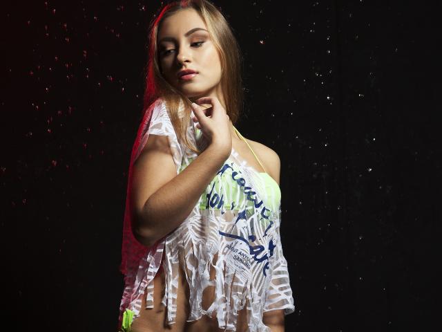 MerindaFoxy mit blonden Haaren, trägt ein durchsichtiges weißes Oberteil mit blauen und grünen Mustern, das ihre kleinen Brüste zeigt, und einen neon-grünen Bikini-Hosen, vor einem dunklen Hintergrund..