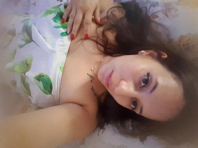 RenattaRosse sdraiata sul letto con i capelli castano ricci, le unghie rosse, un vestito bianco con foglie verdi e un tatuaggio sul petto destro, che guarda la fotocamera con un leggero sorriso..