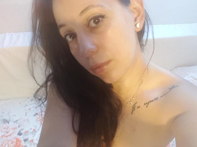 Renatta Rosse mit langen, dunklen Haaren, freiliegenden Schultern, glänzlichen Lippen, kleinen Ohrringen, einem Tattoo an der rechten Schulter, einer Halskette, die in die Kamera schaut, weiches Licht, Bett mit blumigem Bettbezug im Hintergrund..