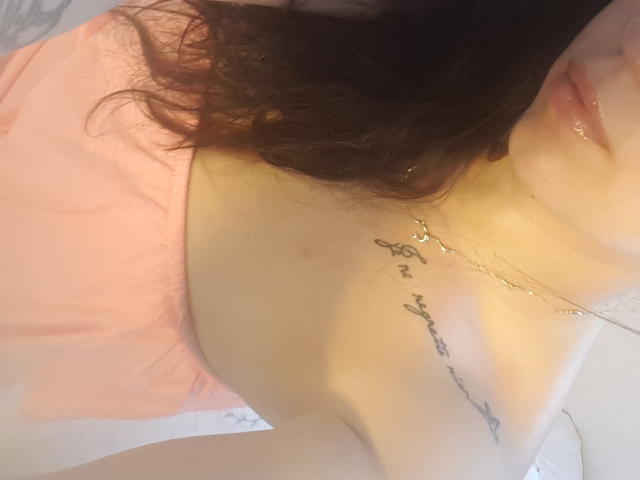 Los senos de RenattaRosse son visibles con una blusa rosa, mostrando un tatuaje en el cuello y el pecho, y lleva un collar. Está acostada, con el pelo castaño ondulado y los labios brillantes..