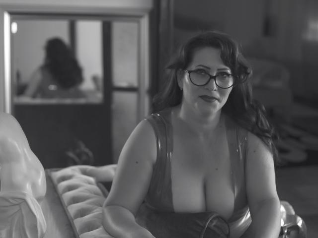 SoniaLaBelle with glasses, medium tits, and dark hair, wearing a shiny dress, sitting on a bed, looking at the camera, in a black-and-white photo.