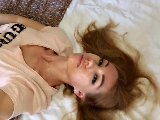 SugarLover sdraiata su un letto, che guarda verso l'alto con occhi verdi, indossa una maglietta rosa, mostrando un seno piccolo, con capelli biondi mossi sparsi, una luce soffusa sul viso..