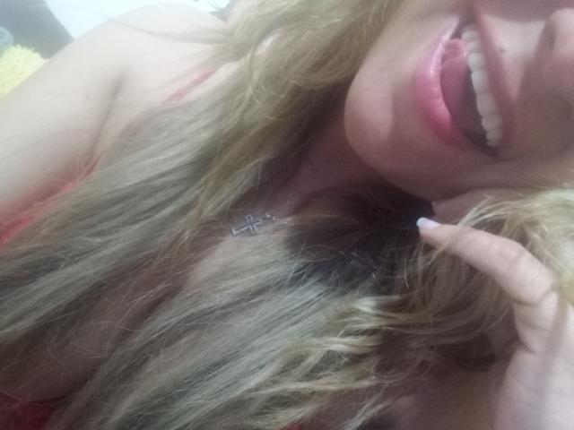 Angelonix with blonde hair, pink lips, and a slight smile, showing teeth, touching her cheek with a finger, wearing a red top, and a cross necklace.