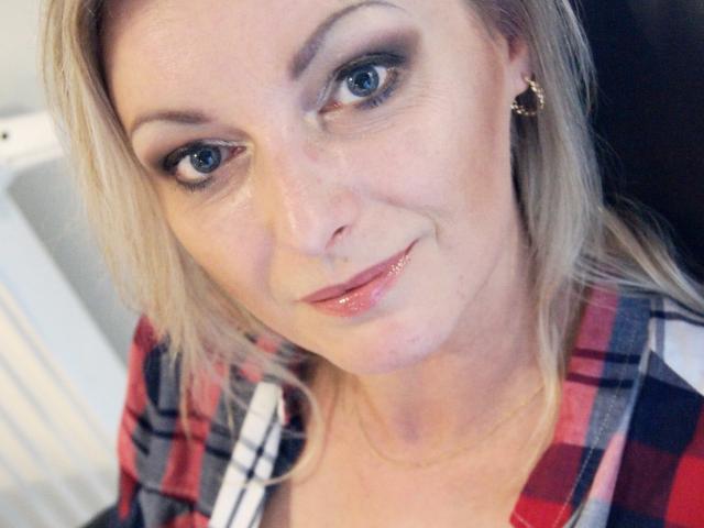 XLovelyVanessa, con cabello rubio, ojos azules y labios rosados brillantes, vestida con una camisa roja de cuadros, tiene una sonrisa encantadora y lleva aretes de aro dorados, en un selfie..