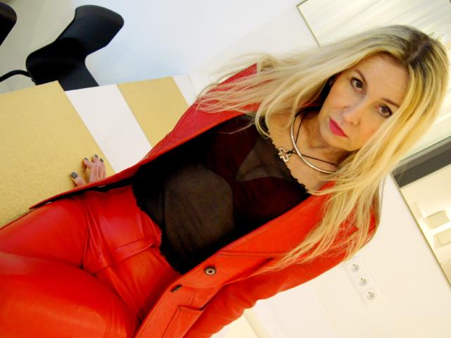 StarCrystal with blonde hair, wearing a red leather jacket, black sheer top, and red pants, has a necklace and purple nails, looking directly at the camera with a neutral expression.