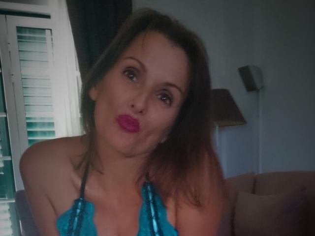 MelindaHottX with pouty pink lips, brown hair, blue halter top, showing her tits, in dimly lit room, looking at camera with slightly tilted head.
