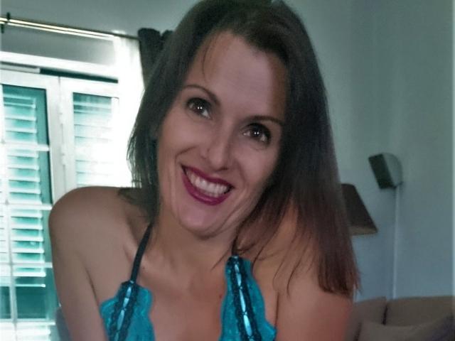 MelindaHottX with a big smile, wearing a blue sequin halter top, showing her tits, brown hair, and pink lipstick, in a bright room with green shutters.