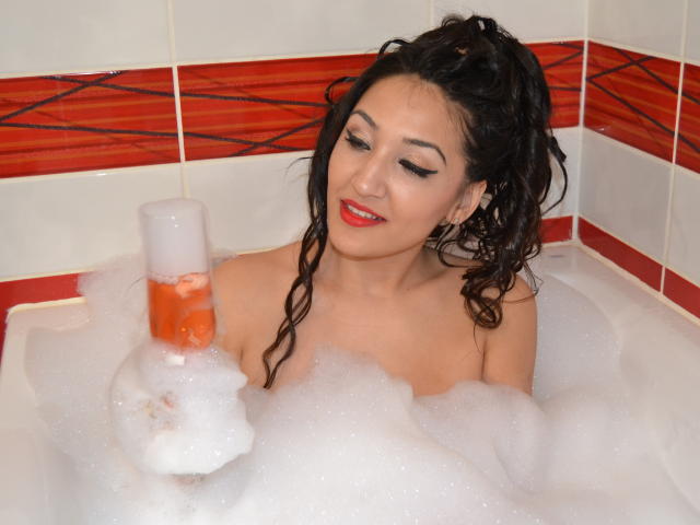 AlesyaNoova with wet black hair in a bubble bath, holding a bottle of orange liquid, smiling with red lipstick, showing her tits, surrounded by white foam.
