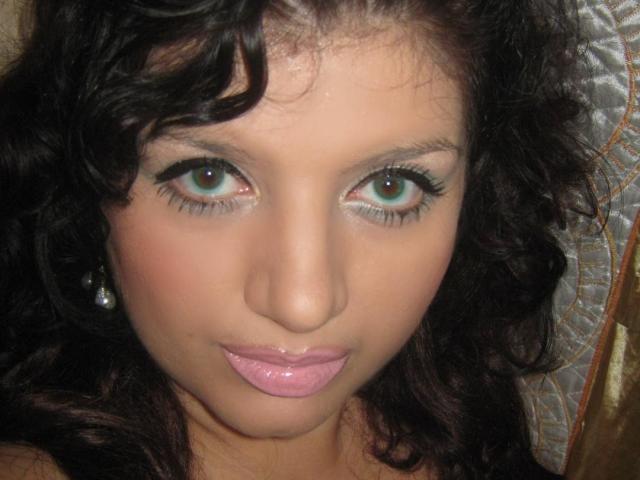 Freyja has curly black hair, green eyes, and glossy pink lips. She's wearing small pearl earrings and has a slightly shiny nose. Her face is centered in the image.
