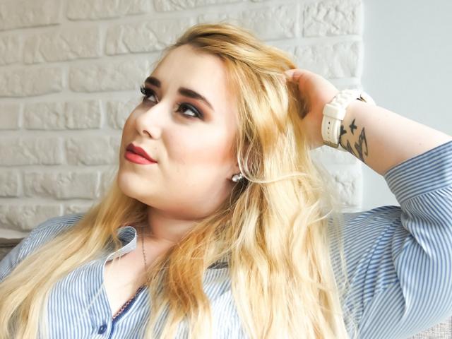 HaileyLush tiene el cabello rubio, lápiz labial rojo y lleva una camisa a rayas con un reloj blanco. Está mirando hacia arriba, con la mano en el pelo, mostrando un tatuaje en su muñeca..