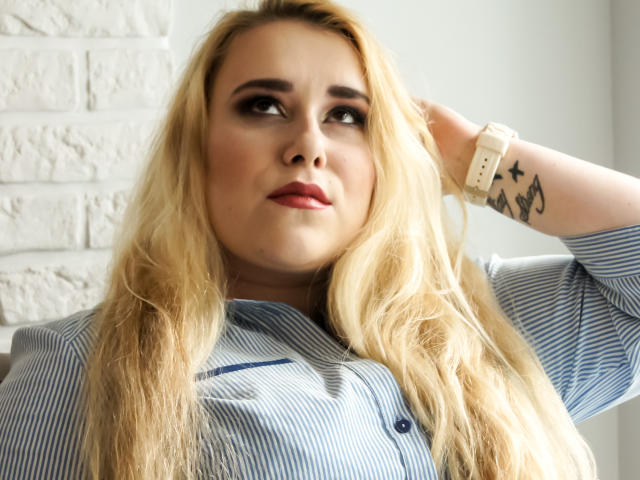 HaileyLush, com cabelos loiros longos, vestindo uma camisa azul listrada, olhando para cima com uma expressão ligeiramente confusa, com a mão no cabelo, parede de tijolos brancos ao fundo, tatuagem no pulso..