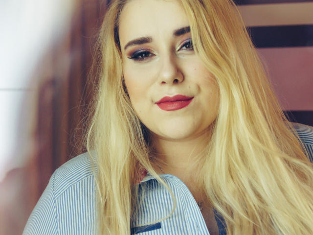 HaileyLush, met blond haar, blauwe ogen en rode lippenstift, draagt een blauw gestreepte blouse en kijkt iets opzij met een subtiele glimlach. Haar borsten zijn niet zichtbaar..