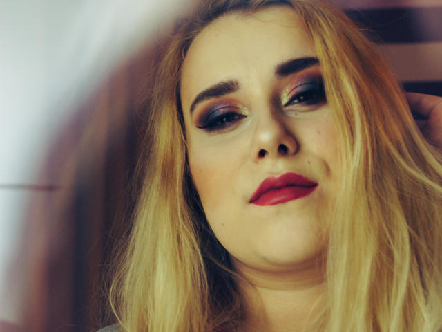 HaileyLush has blonde hair, red lipstick, and smoky eye makeup, looking slightly annoyed with a small mole on her right cheek. Her tits are partially visible.