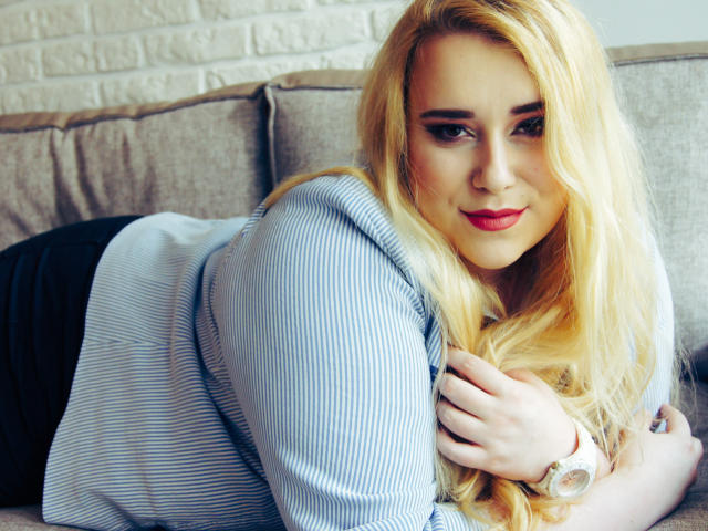 HaileyLush mit langen blonden Haaren, rotem Lippenstift und einem blau gestreiften Hemd, liegt auf einem Sofa, schaut in die Kamera, ihre Hand liegt auf ihrer Brust, und sie lächelt leicht..