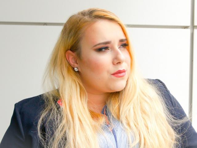HaileyLush tem cabelo loiro, olhos azuis, batom rosa e está vestindo um blazer preto sobre uma camisa listrada. Ela está olhando para o lado com uma expressão levemente emburrada..
