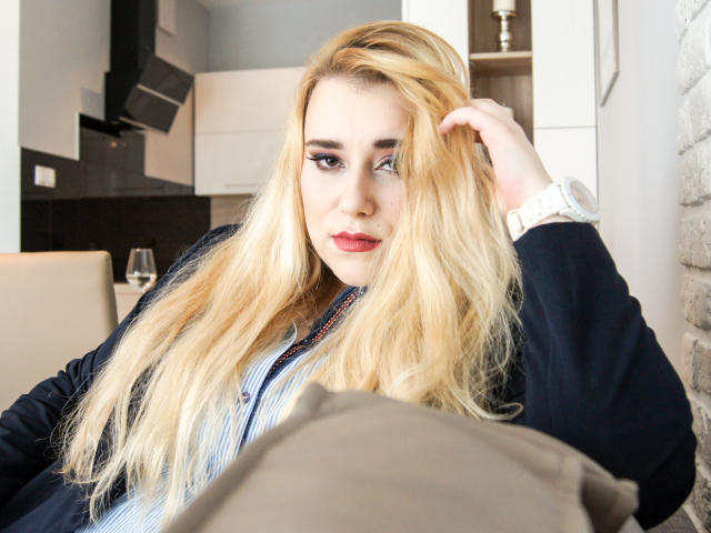 HaileyLush, loira, com cabelo comprido, batom vermelho e delineador, vestindo uma camisa listrada de azul e um casaco preto, toca no cabelo enquanto olha para a câmera. Ela está usando um relógio branco..