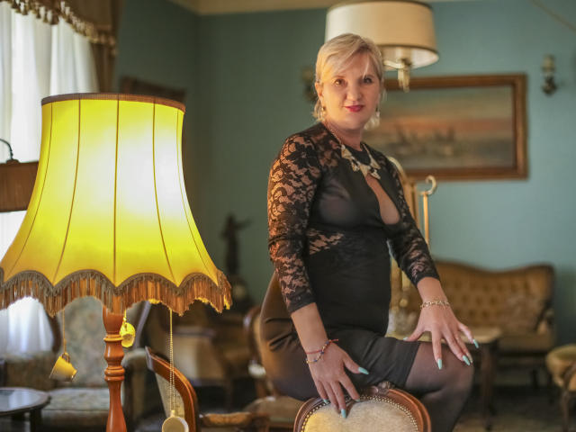 ExperiencedAlana with blonde hair, wearing a black lace dress showing her tits, sitting on a chair, hands on her thighs, in a room with a yellow lamp.
