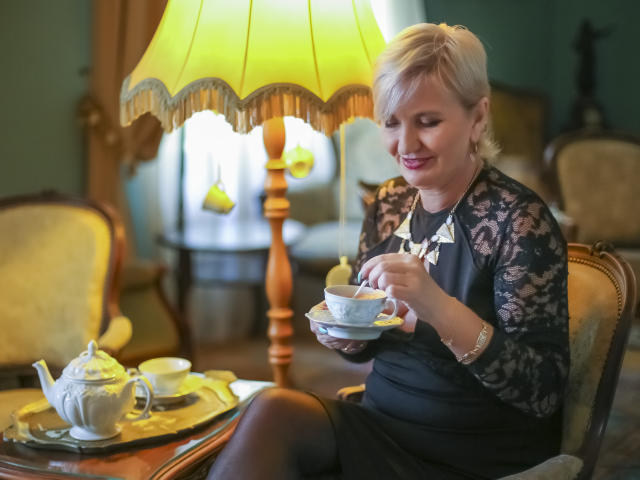 Alana, experiente, com cabelo loiro curto, vestindo uma blusa preta de renda, tomando um gole de uma xícara de chá azul, sentada em uma sala elegante com um abajur amarelo, segurando uma bandeja de chá..