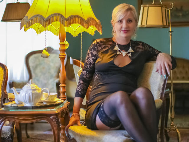 ExperiencedAlana, a blonde woman with large boobs and a curvy ass, sits on a chair wearing a black lace dress, garter, and stockings, with a yellow lamp beside her.