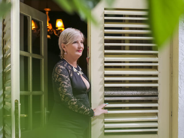 ErvarenAlana, met kort blond haar, een zwarte kanten jurk en een ketting, staat in een raam met een groene lijst en laat haar grote borsten zien, ze glimlacht en houdt aan een luik vast, met groene bladeren op de voorgrond..