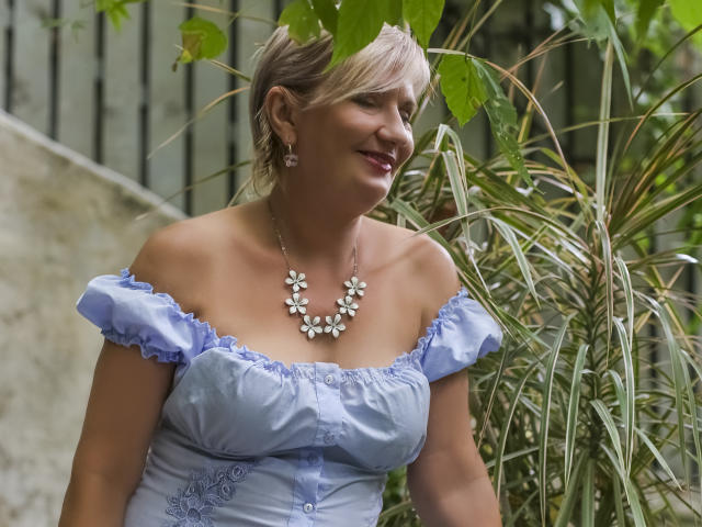 ExperiencedAlana with blonde hair, off-shoulder blue dress, floral necklace, smiling, standing near green plants, showing her tits, wearing earrings, looking to the side.