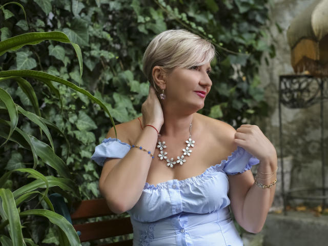 ExperiencedAlana with short blonde hair, wearing a light blue off-shoulder dress, floral necklace, and red bracelet, touching her neck, surrounded by green plants. She's looking to the right.
