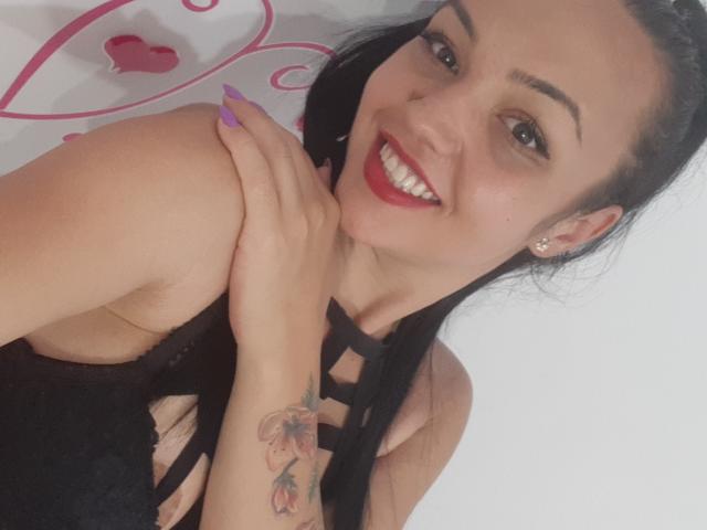 FunBrunettee with black hair, red lipstick, and purple nails, smiling at the camera, showing a tattoo on her arm, wearing a black lace top, with visible tits.