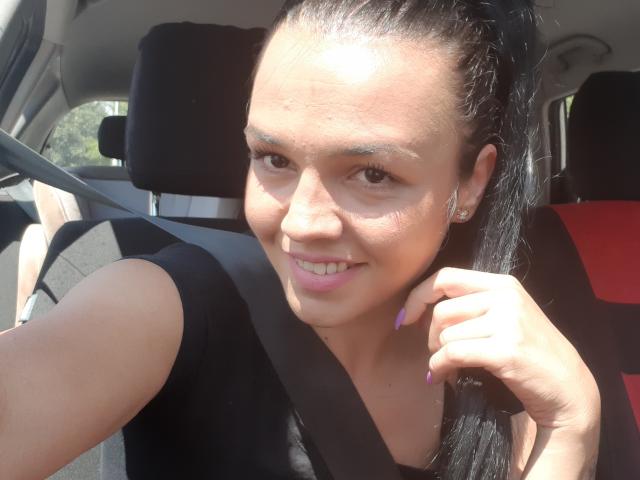 FunBrunettee with dark hair in a ponytail, wearing a black shirt, smiling at the camera, seatbelt across chest, purple nails, and slight freckles on her face.