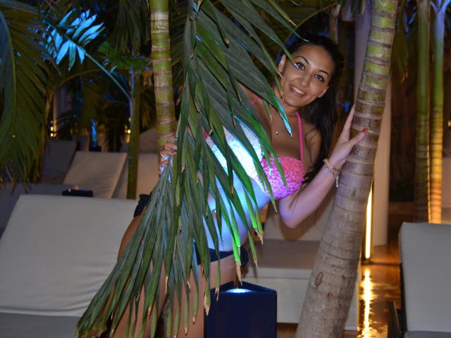 AlesyaNoova mit mittelgroßer Brust, trägt ein pinkes Spitzenoberteil, schwarze Shorts und eine Sonnenbrille, versteckt sich hinter einem Palmenbaum, lächelt und hält den Baum mit ihrer rechten Hand..