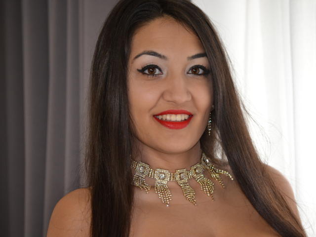 AlesyaNoova has long brown hair, red lipstick, and a gold necklace with dangling jewels. She's smiling with brown eyes, wearing red lipstick, and showing her bare shoulders.