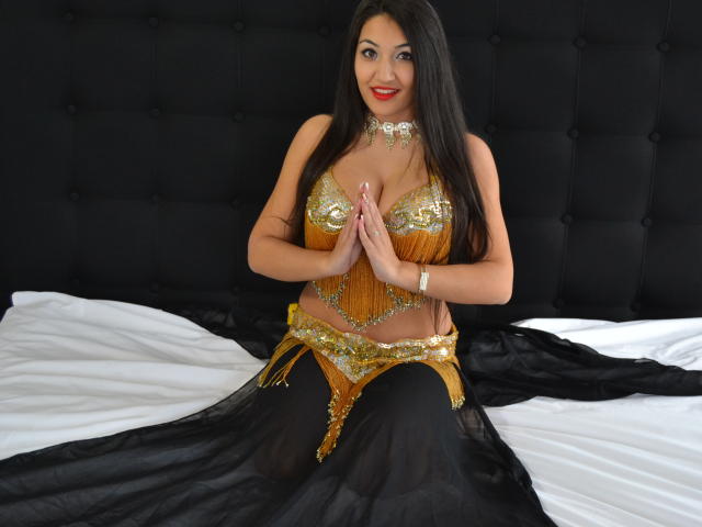AlesyaNoova, con cabello negro largo, lápiz labial rojo, un traje de bailarina del vientre dorado y pantalones harem negros, sentada en una cama, con las manos juntas, sonriendo, mostrando sus pechos y abdomen brillantes..