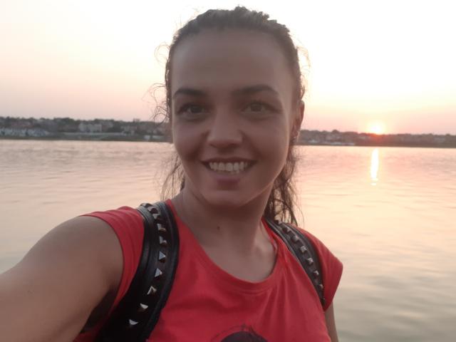FunBrunettee with puffy lips, smiling, wearing a red shirt, black backpack with spikes, brown hair in a bun, standing by a sunset over a calm water body.