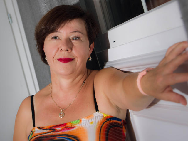 AdeleLoveEx, middle-aged woman with short brown hair, red lipstick, wearing a colorful dress, black straps, gold necklace, reaching out, smiling, white door in background, pink wristband.
