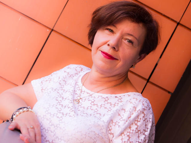 AdeleLoveEx with short brown hair, red lipstick, white lace top, necklace, bracelets, smiling, light skin, slightly chubby, sitting against orange wall, looking up, cute, confident.
