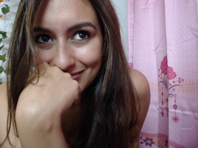 Samantha Belle, con cabello castaño largo, sonriendo, ojos marrones, y su mano cerca de su rostro sonriente y de nariz rosada. Muestra sus hombros desnudos. Es linda y tiene un aspecto atractivo..