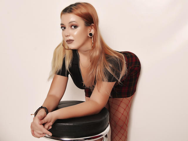 AyannaAngel, avec ses cheveux dégradés blonds et roses, un haut noir, une jupe à carreaux rouges, des bas résille, appuyée sur un tabouret noir, portant des boucles d'oreilles noires, regardant l'appareil photo avec les lèvres légèrement entrouvertes..