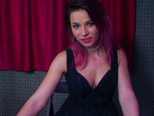 AliciaKristal with pink-dyed hair, wearing a black lace dress with deep cleavage, silver choker, and red lipstick, sitting on a chair, smiling at the camera.