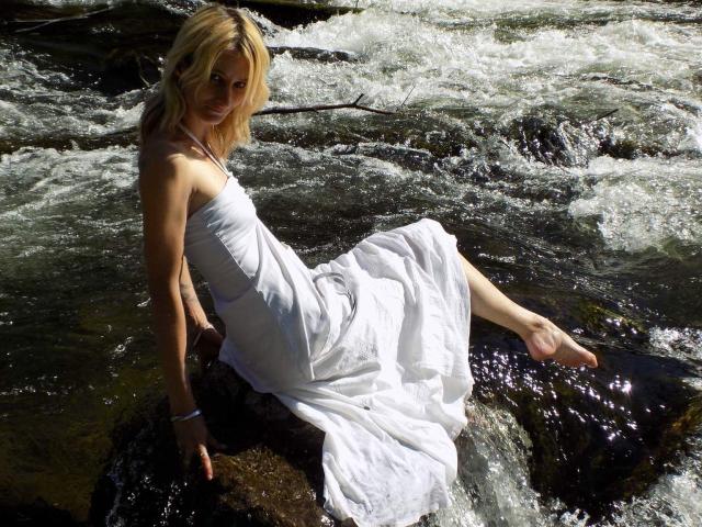 SauterelleSexy com cabelo loiro, vestido branco, sentada em pedras em um riacho, seus pés descalços na água, sua bunda ligeiramente visível, vestido molhado, olhando para a câmera..
