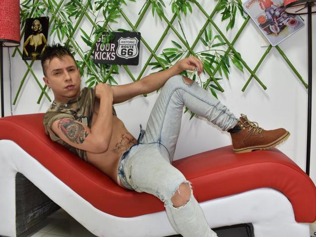 LenyHotCock, un homme mince avec des tatouages, portant une chemise marron, un jean déchiré, des bottes marron, allongé sur un transat rouge et blanc, tenant des feuilles vertes, visage sérieux, cheveux bruns courts, abdominaux visibles..