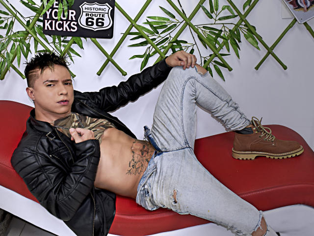 LenyHotCock, mit kurzen schwarzen Haaren, einer Lederjacke, zerrissenen Jeans, braunen Stiefeln und tätowierten Bauchmuskeln, sitzt entspannt auf einer roten Bank und präsentiert eine selbstbewusste, sexy und gelassene Pose..
