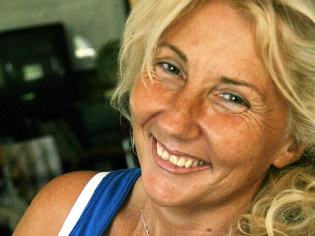 SusanaBlondeHot a les cheveux blonds et bouclés, des yeux bleus, et un visage souriant avec des rides, elle porte un haut bleu, montre ses dents et a une expression légèrement amusante. Ses seins ne sont pas visibles..