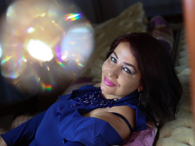 LexyRose mit dunkelroten Haaren, einem blauen Kleid und pinkem Lippenstift, liegt auf einem Bett und betrachtet eine farbenfrohe, leuchtende Kugel. Dabei ist ihre nackte Schulter und ein dezenter Ausschnitt zu sehen..