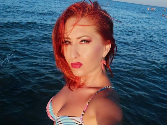 IvyFox, con cabello rojo, vestida con un bikini a rayas, mostrando sus pechos en el agua, con lápiz labial rojo y aretes rosas, mirando a la cámara con una expresión ligeramente molesta..