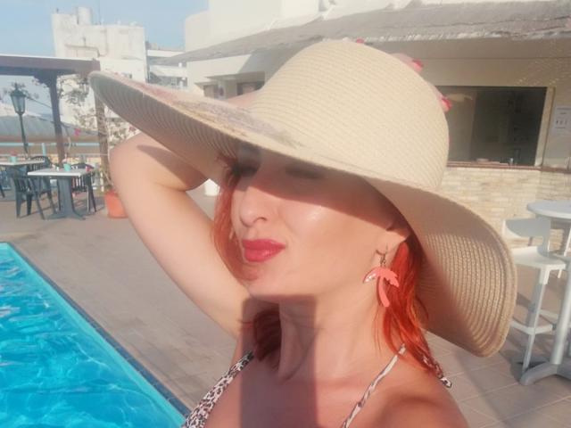 IvyFox with red hair, wearing a wide-brimmed beige hat, pink earrings, and a leopard print bikini, puckering her lips by a blue pool. Her tits are visible.