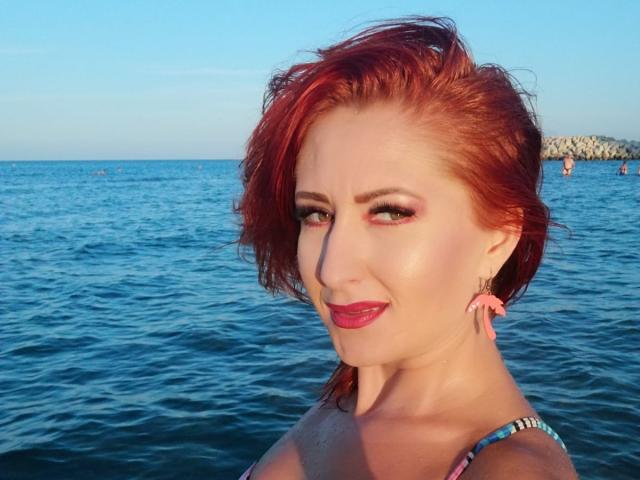 IvyFox with red hair, pink earrings, and red lipstick, showing off her tits in a blue bikini, smiling at the camera, standing in the ocean.