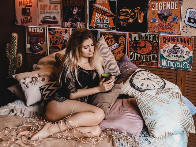 ShyNaomi, with long blonde hair, sits on a bed in black lace lingerie, holding a green cup, her bare ass and tits visible, surrounded by pillows and vintage posters.