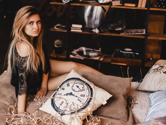 ShyNaomi met lang blond haar, een zwarte kanten top en mouwen die van de schouder vallen, zittend op een bed met een kussen in de vorm van een klok, kijkt ze over haar schouder, waarbij haar achterkant lichtelijk zichtbaar is..
