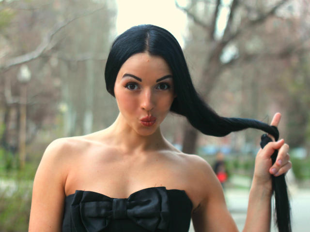RoseCream, con el pelo largo y negro, haciendo pucheros, sujetándose el pelo con una mano, vestida con un vestido negro sin tirantes con un lazo, de pie en un parque, mostrando sus pechos..