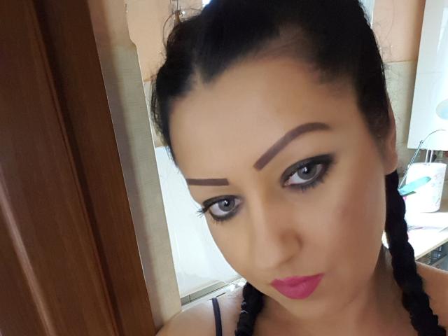 BeauxYeuxx a des sourcils foncés, un eye-liner noir et un rouge à lèvres rose. Elle a des tresses noires, des yeux bleus et une expression sérieuse, avec le visage légèrement incliné. Ses seins sont partiellement visibles..